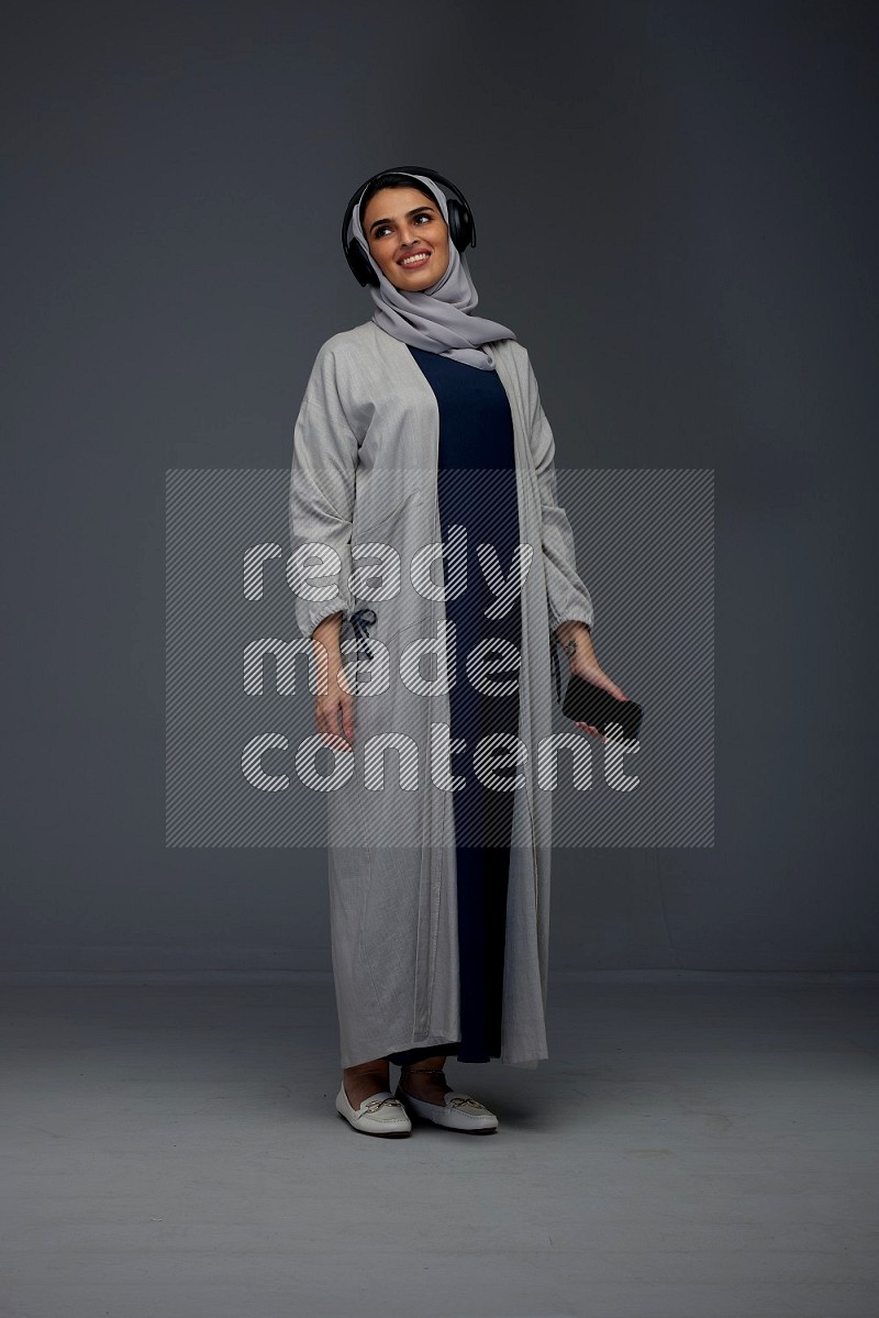 A Saudi woman wearing a light gray Abaya and head scarf standing and listening to music on a grey background