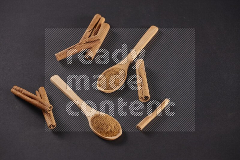 Two wooden spoons full of cinnamon powder with cinnamon sticks on black background