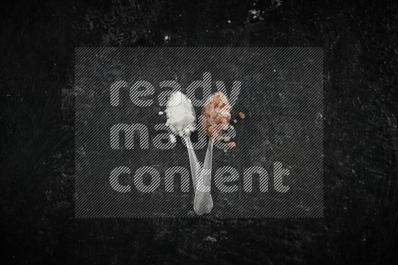 2 metal spoons filled with white salt and pink himalayan salt on black background