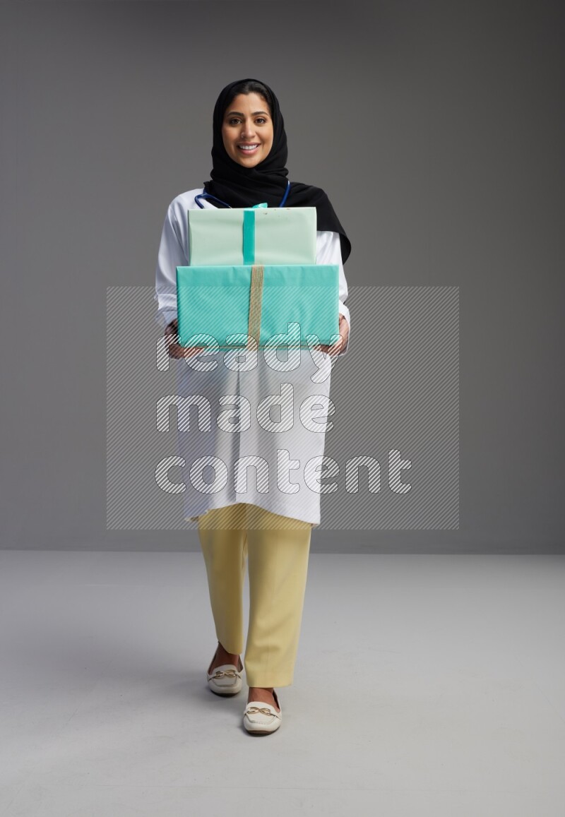 Saudi woman wearing lab coat with stethoscope standing holding gift box on Gray background
