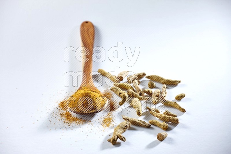 A wooden ladle full of turmeric powder and dried turmeric fingers beside it on white flooring