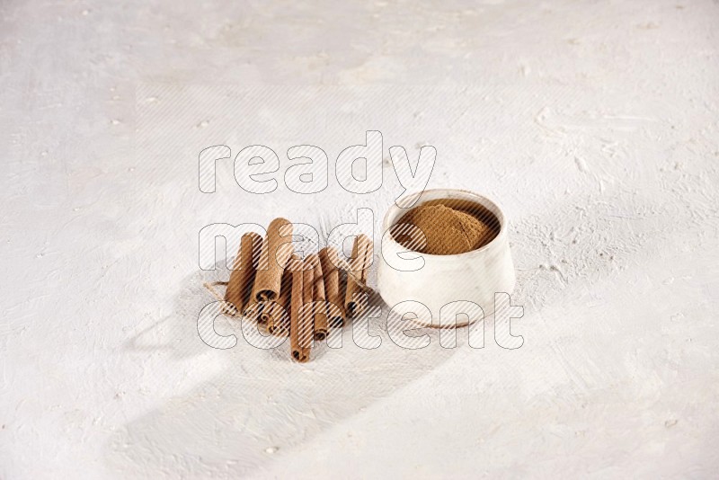 Cinnamon sticks stacked beside a beige bowl full of cinnamon powder on white background