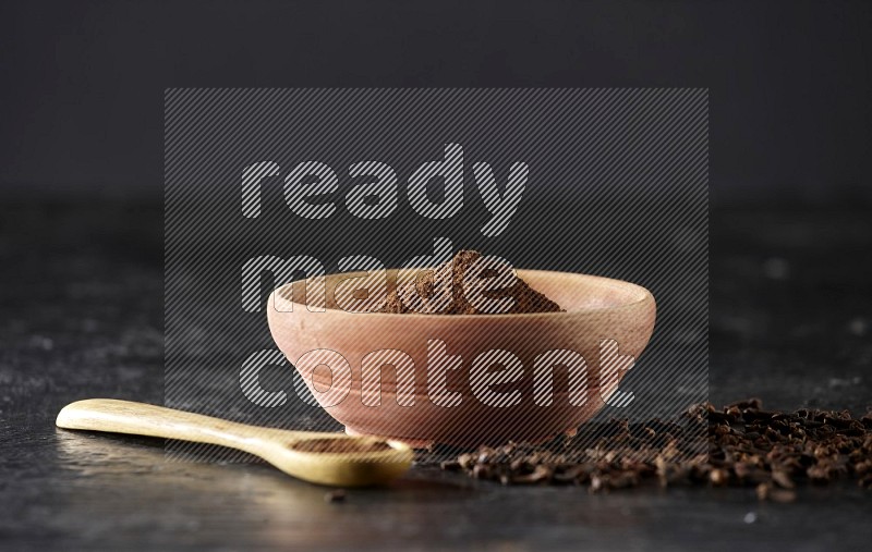 A wooden bowl and wooden spoon full of cloves powder with spreaded cloves on textured black flooring