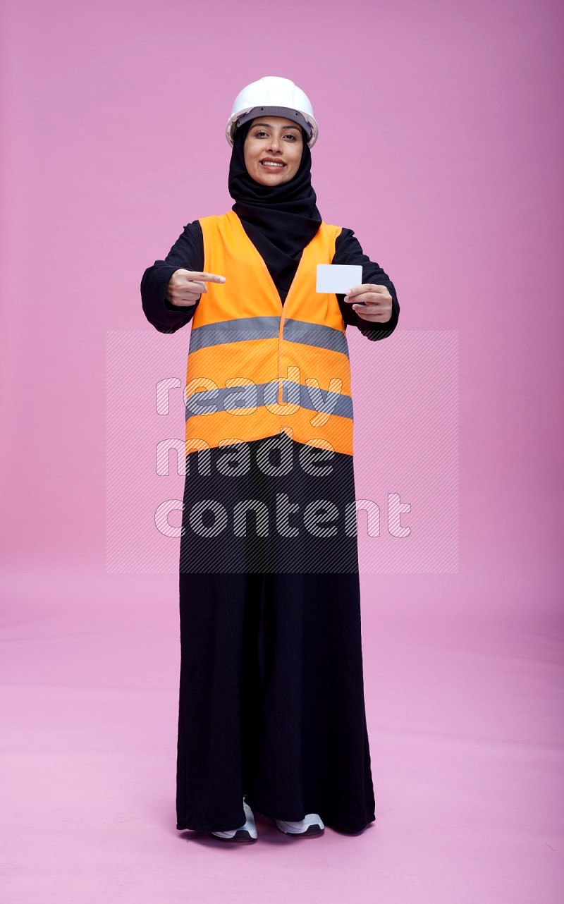 Saudi woman wearing Abaya with engineer vest and helmet standing holding ATM card on pink background