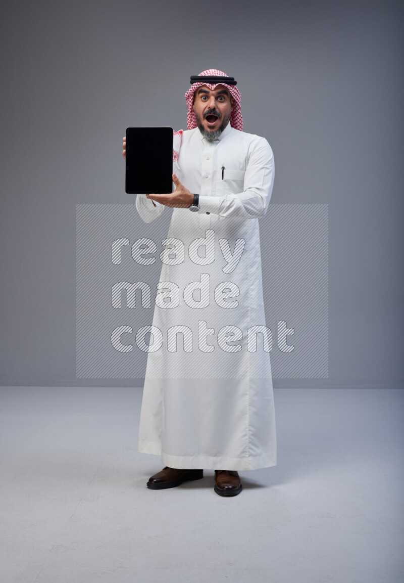 Saudi man Wearing Thob and red Shomag standing showing tablet to camera on Gray background