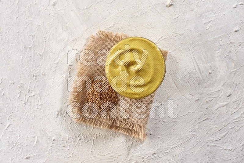 A glass bowl full of mustard paste set on a burlap piece with some mustard seeds spread on a textured white flooring