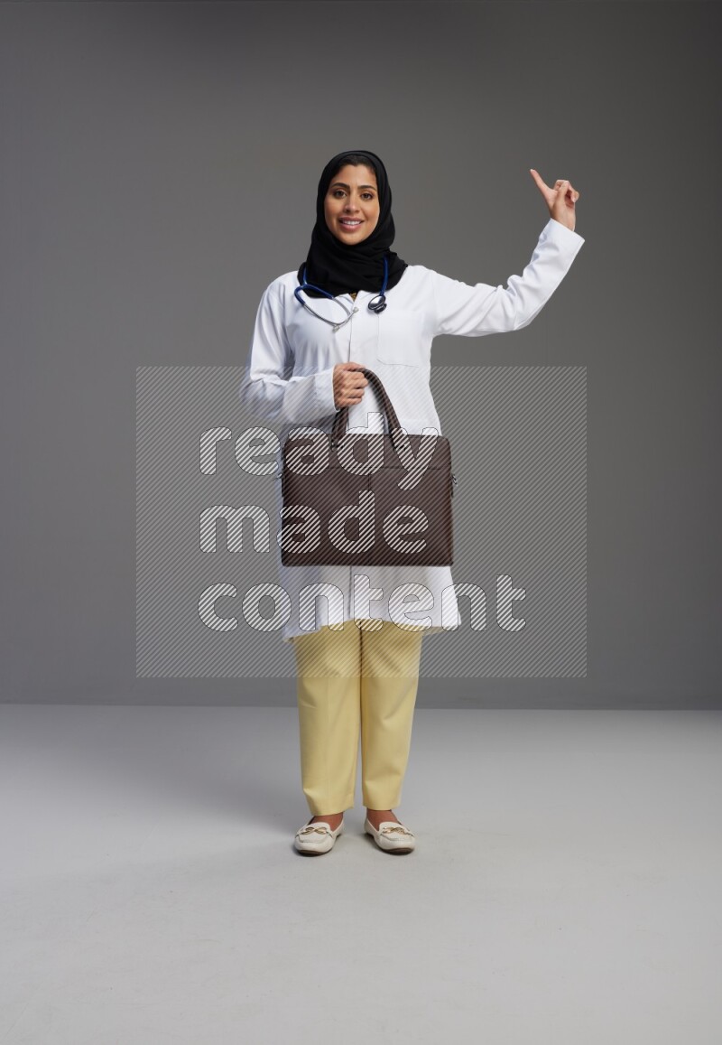Saudi woman wearing lab coat with stethoscope standing holding bag on Gray background