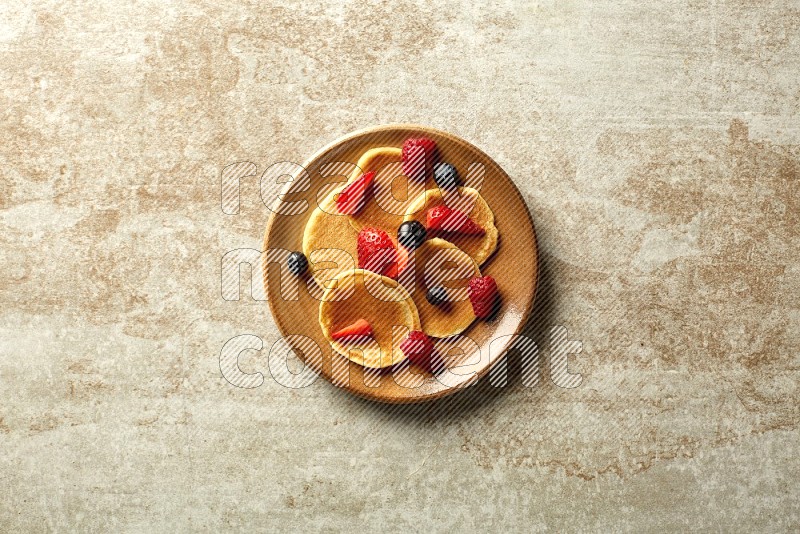 Five stacked mixed berries mini pancakes in a brown plate on beige background