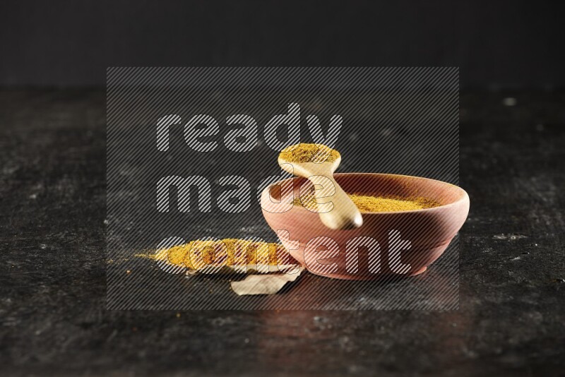 A wooden bowl and a wooden spoon full of turmeric powder on textured black flooring