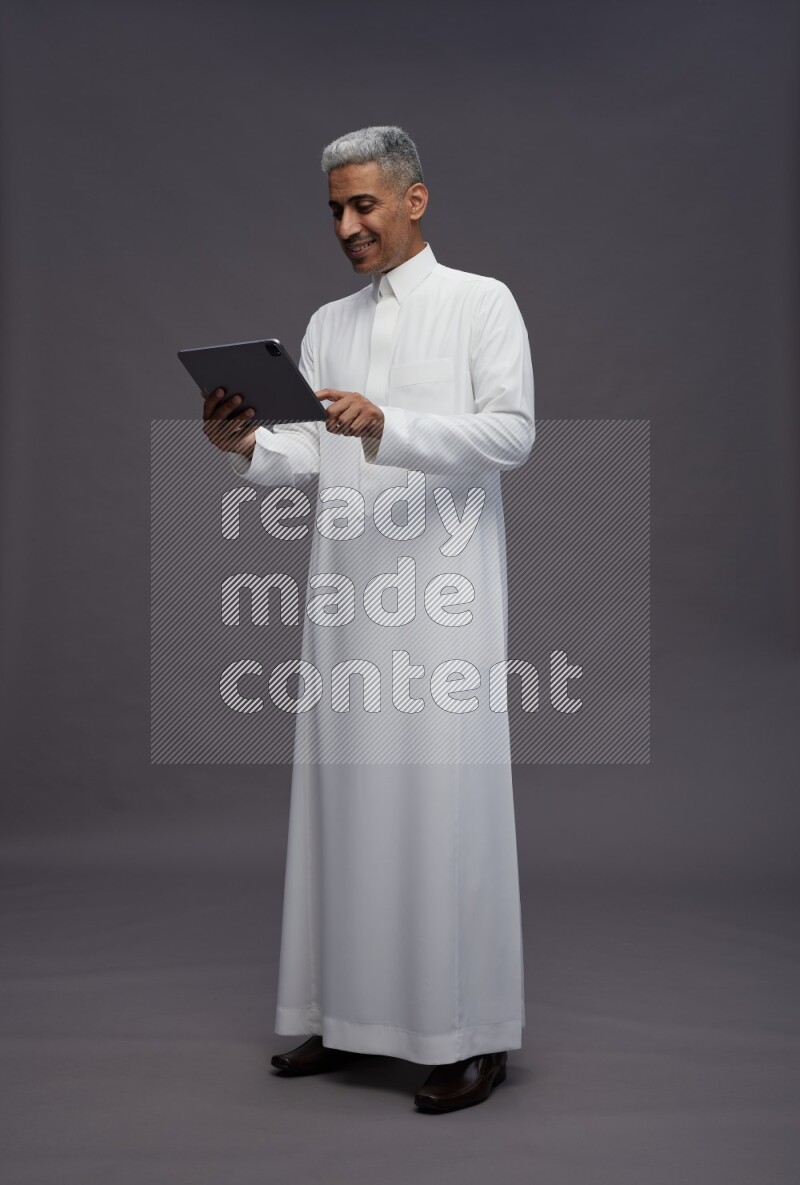 Saudi man wearing thob standing working on tablet on gray background