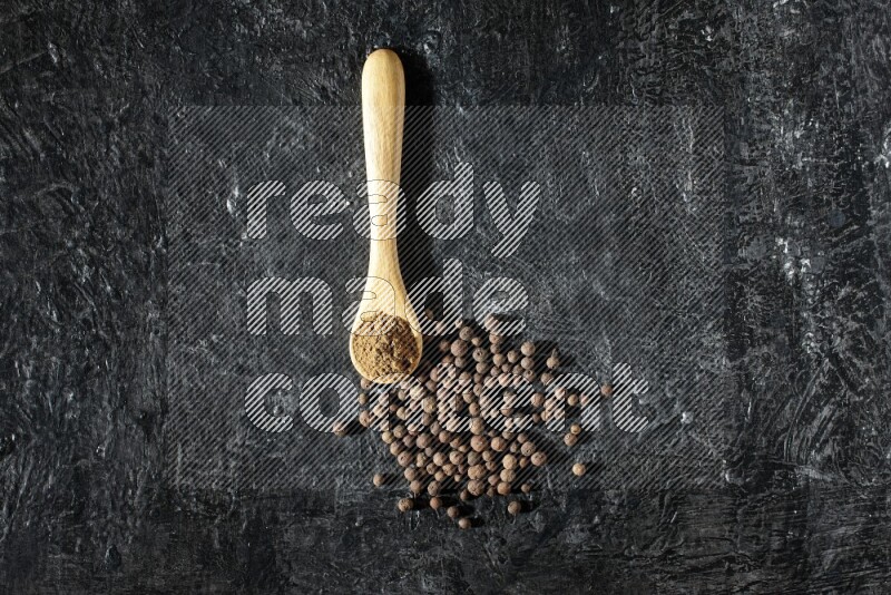 A wooden spoon full of allspice powder and whole balls spreaded on a textured black flooring