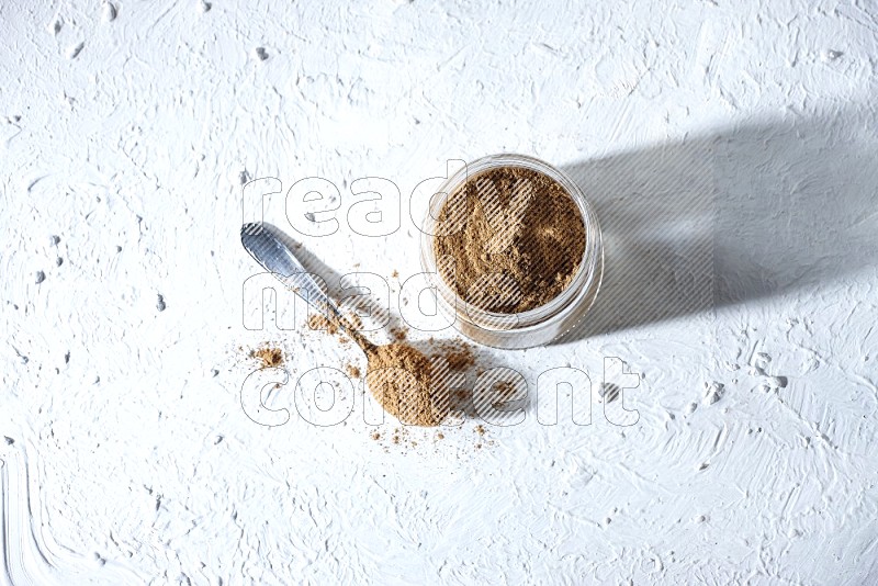 A glass jar and a metal spoon full of allspice powder on a textured white flooring