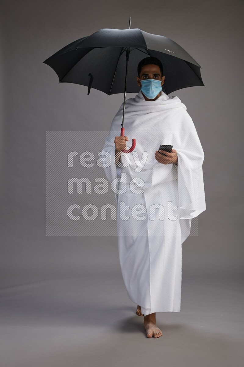A man wearing Ehram and mask Standing holding umbrella on gray background on gray background