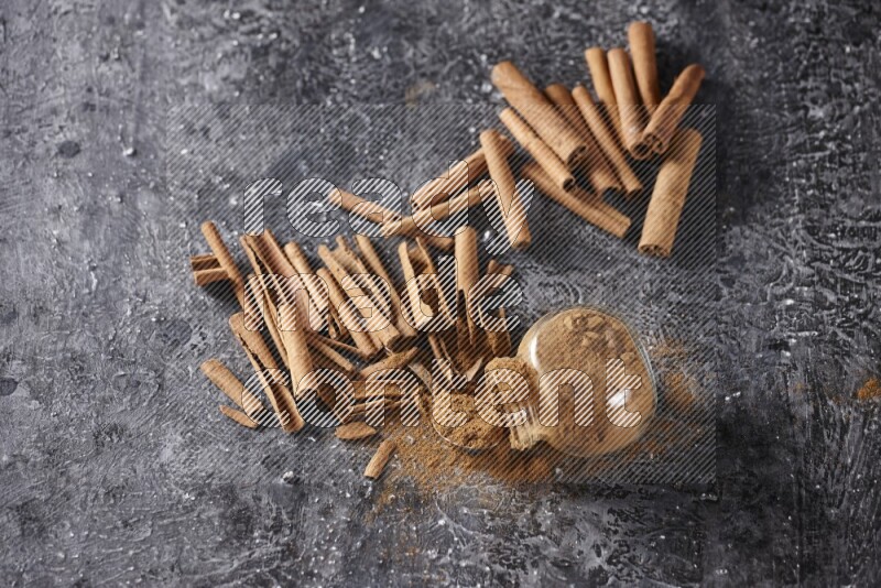 Herbal glass jar full cinnamon powder flipped and a metal spoon full of powder surrounded by cinnamon sticks on textured black background in different angles