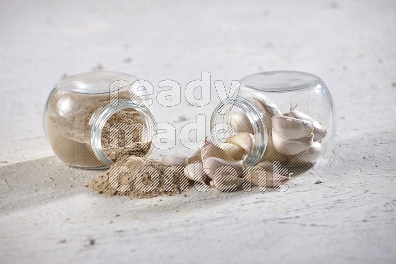 2 glass spice jars full of garlic cloves and powder flipped and their garlic came out on a textured white flooring in different angles