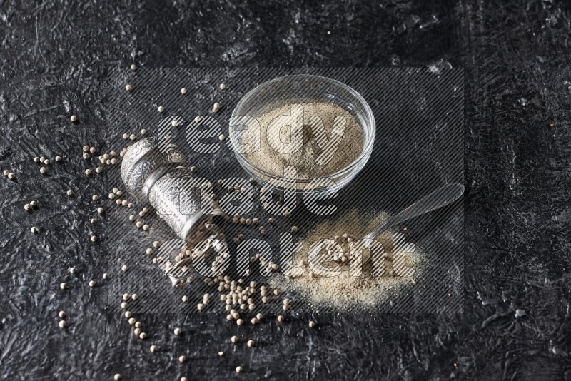 A glass bowl full of white pepper powder with pepper beads, a metal grinder and a metal spoon on textured black flooring