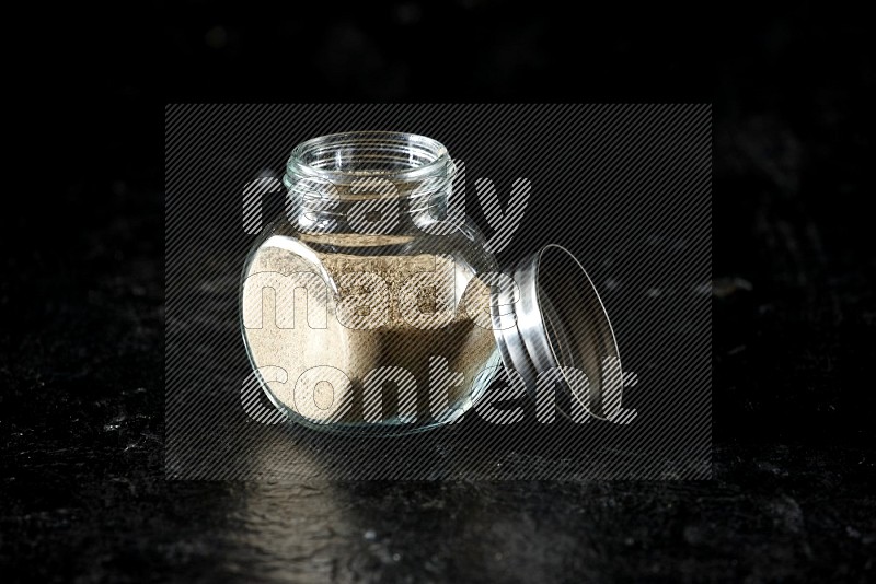 A glass spice jar full of cardamom powder on textured black flooring