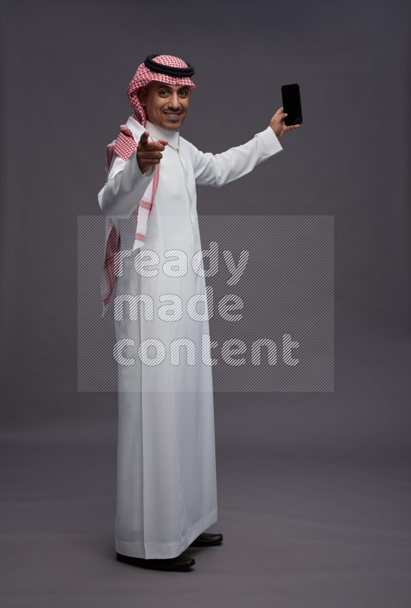 Saudi man wearing thob and shomag standing taking selfie on gray background
