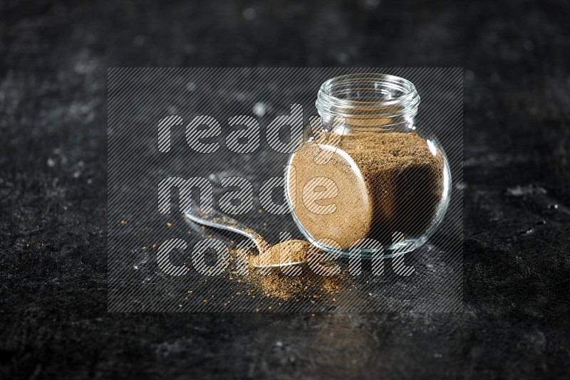 A glass spice jar and a metal spoon full of cumin powder on a textured black flooring