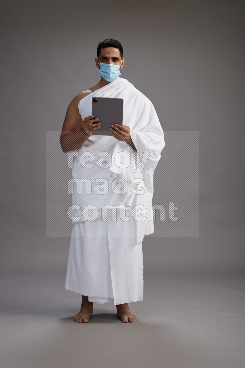 A man wearing Ehram with face mask Standing holding a tablet on gray background