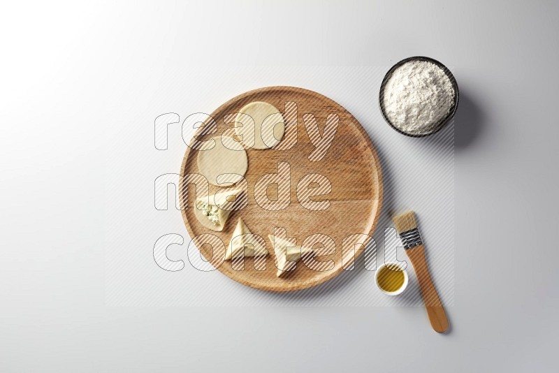 two closed sambosas and one open sambosa filled with cheese while flour, and oil with oil brush aside in a wooden dish on a white background