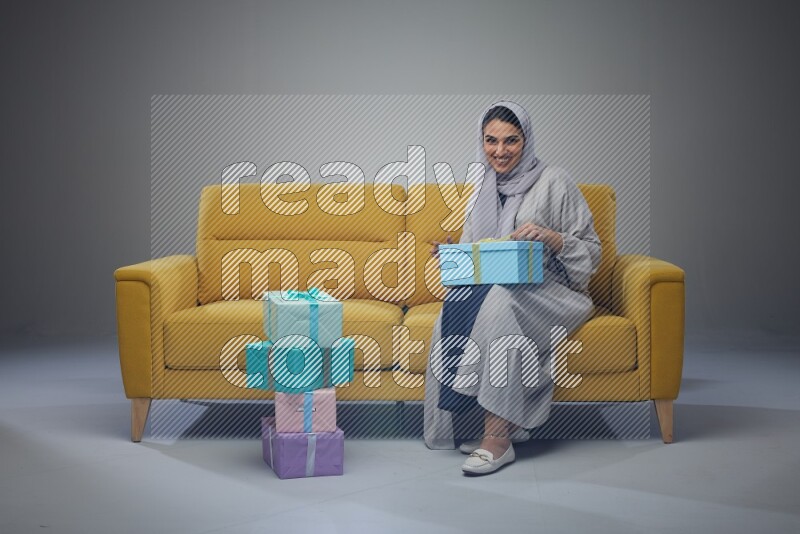 A Saudi woman wearing a light gray Abaya and head scarf sitting on a yellow sofa holding a gift box beside multi color gift boxes eye level on a grey background