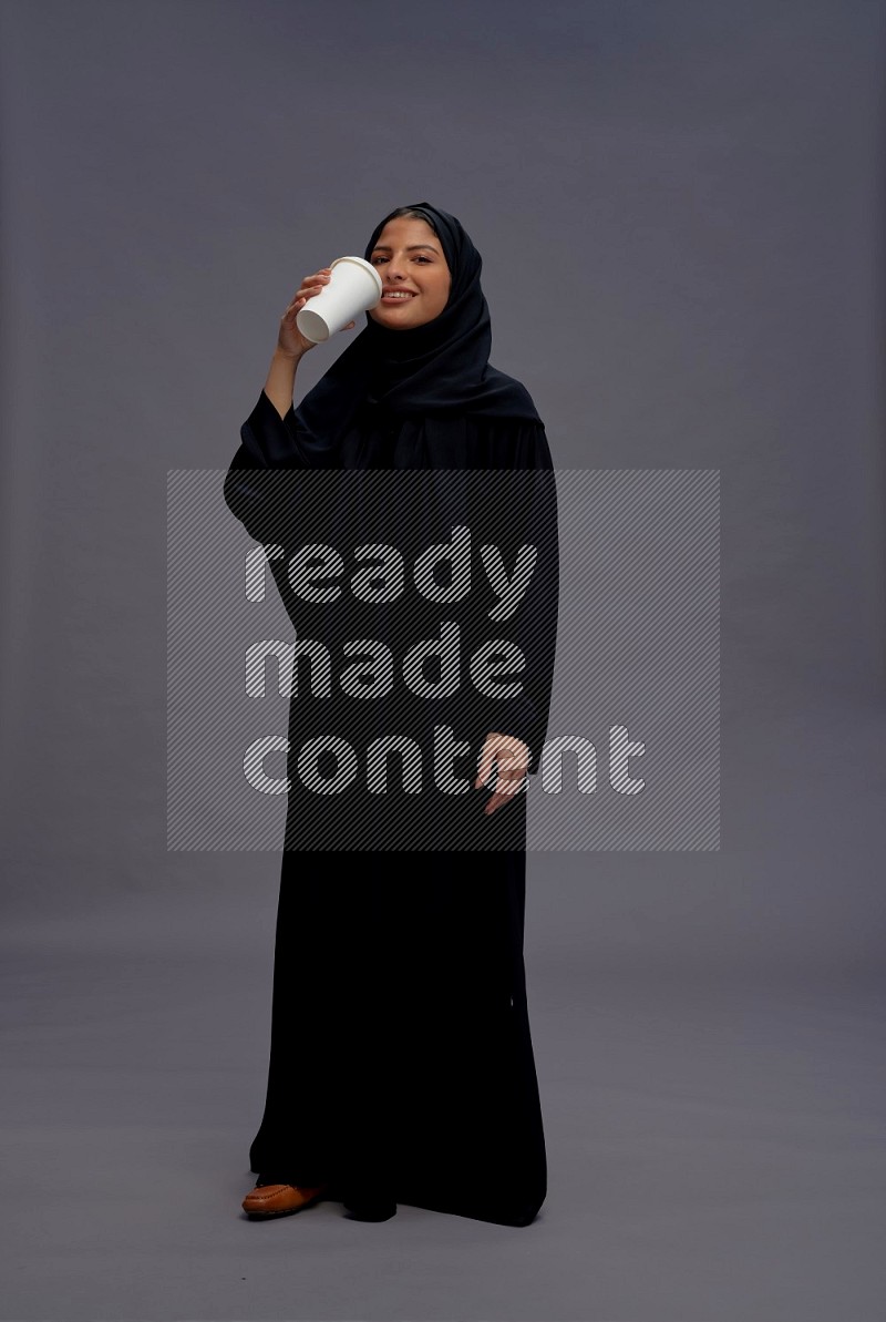 Saudi woman wearing Abaya standing holding paper cup on gray background