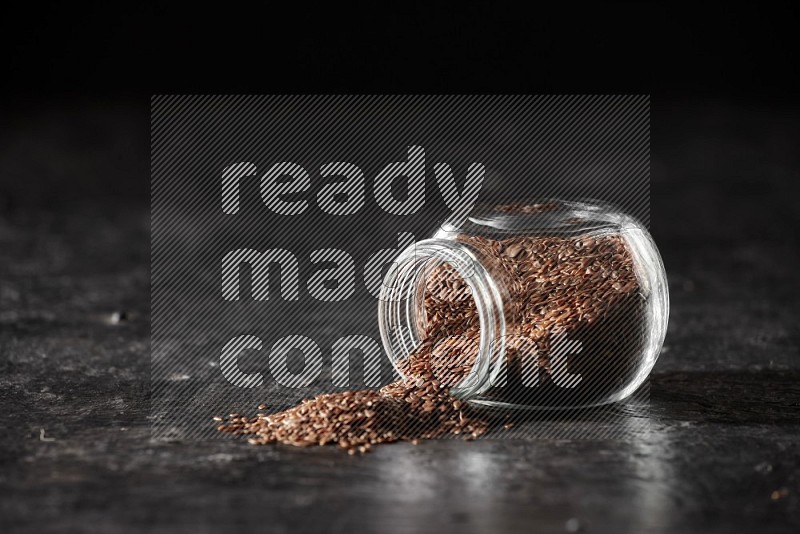 A glass spice jar full of flax flipped and seeds spreaded out on a textured black flooring in different angles