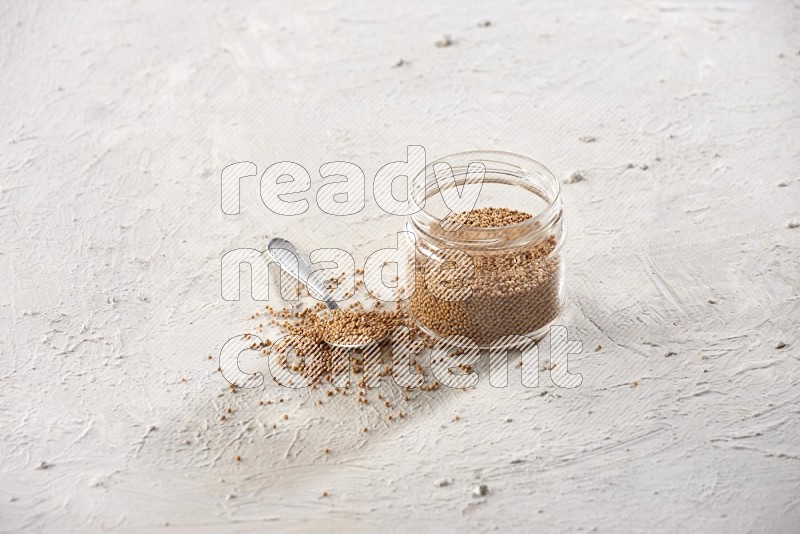 A glass jar and a metal spoon full of mustard seeds on a textured white flooring in different angles
