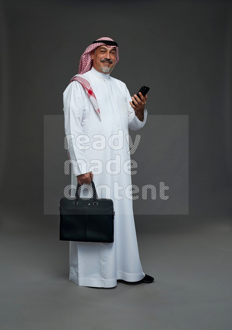 Saudi man with shomag Standing holding bag and phone on gray background