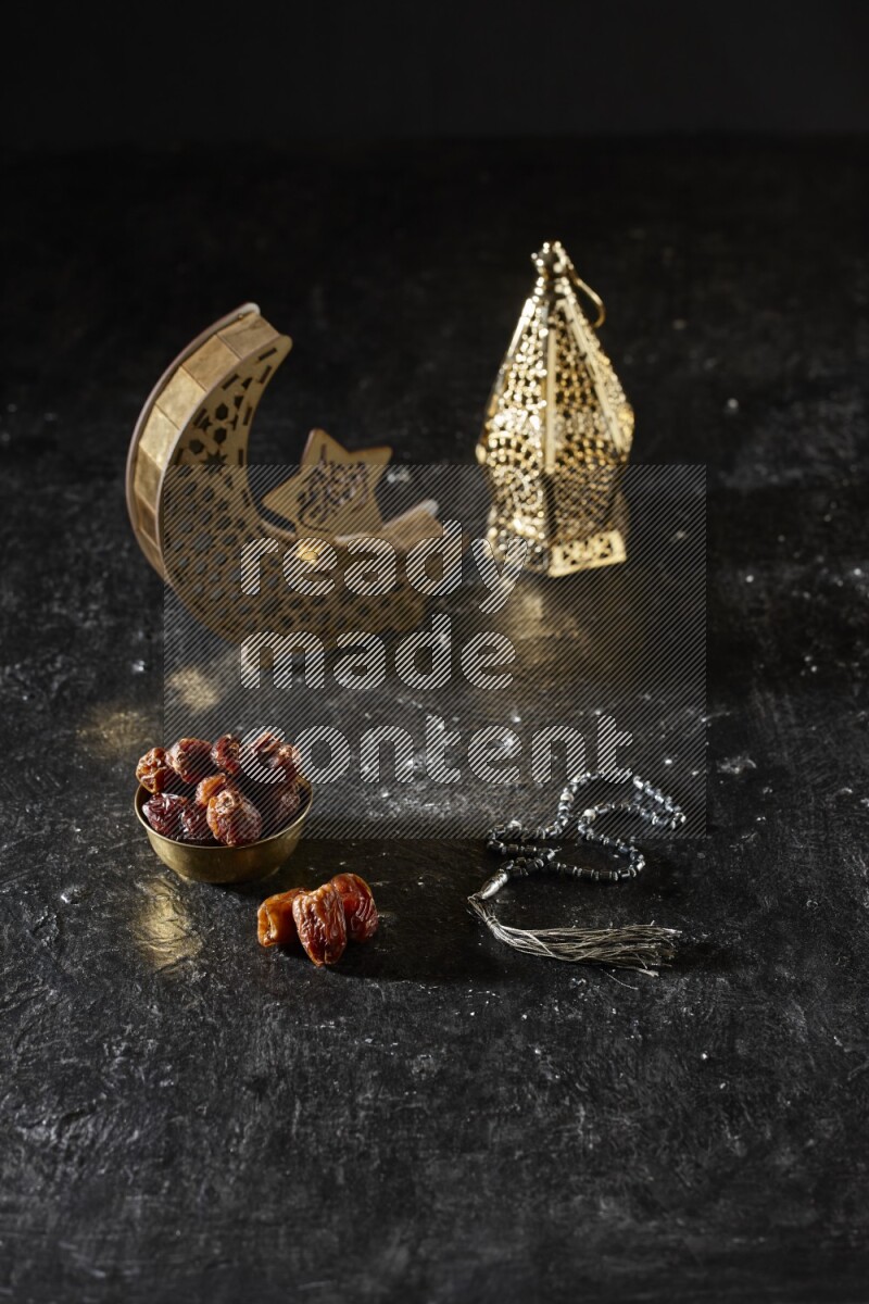 Dates in a metal bowl with prayer beads beside golden lanterns in a dark setup