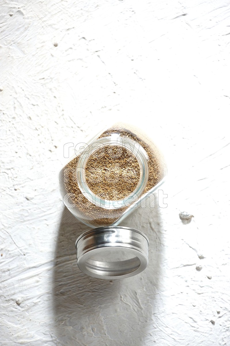 A glass spice jar full of cumin powder on textured white flooring