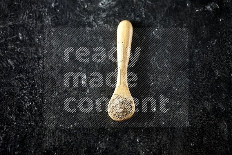 A wooden spoon full of cardamom powder on textured black flooring