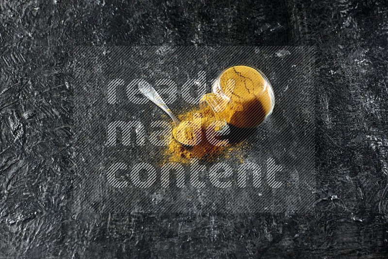 A flipped glass spice jar and a metal spoon full of turmeric powder and powder spilled out of it on textured black flooring