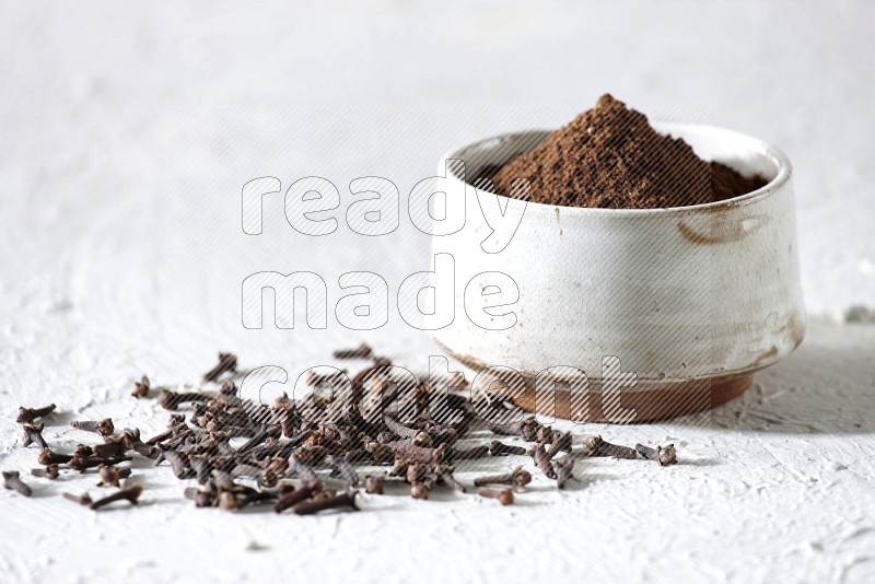 A beige ceramic bowl full of cloves powder and whole cloves on a white flooring