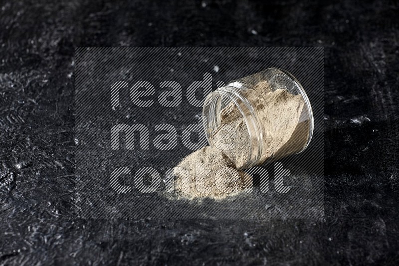 A flipped glass jar full of white pepper powder with spilled powder on textured black flooring