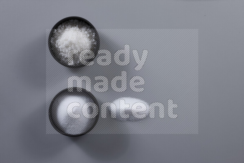2 pottery bowls one is filled with table salt and the other with coarse sea salt on grey background