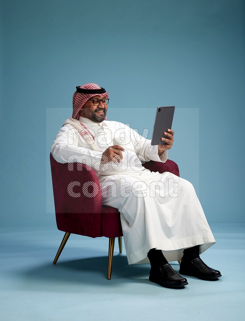 Saudi Man with shimag sitting on chair working on tablet on blue background