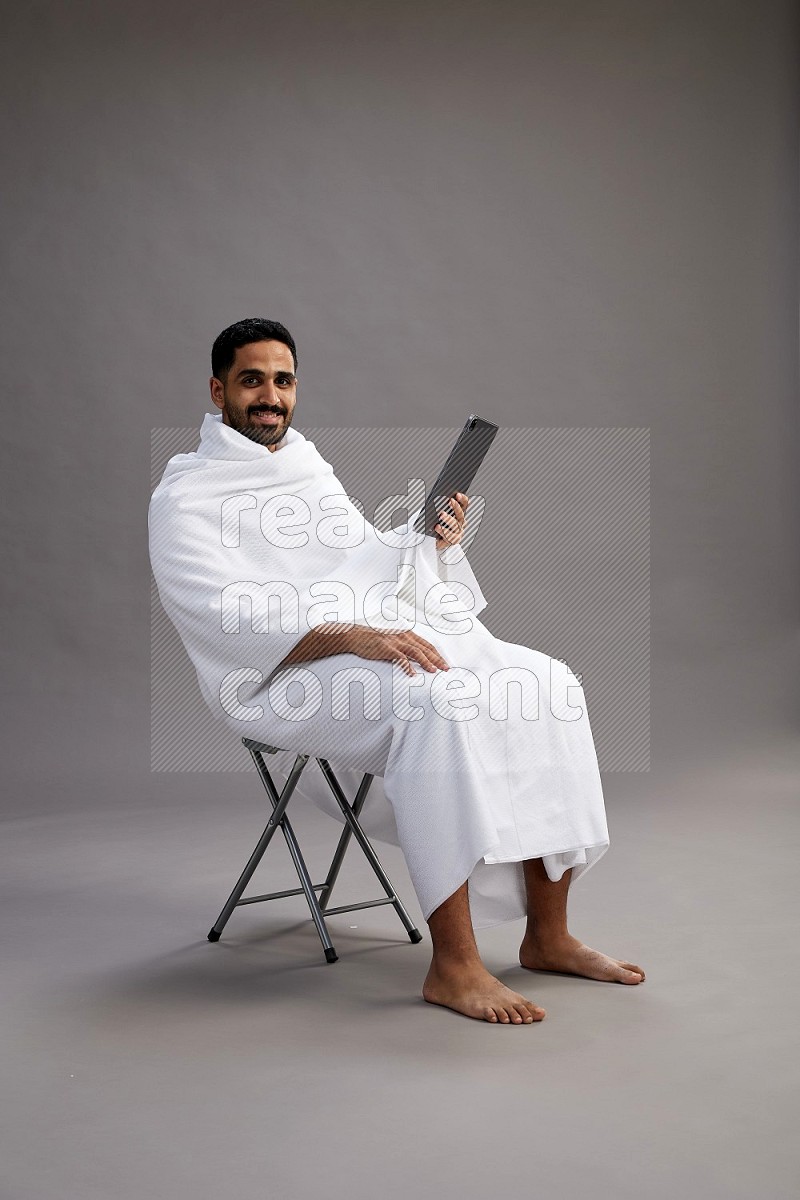 A man wearing Ehram sitting on chair holding a tablet on gray background