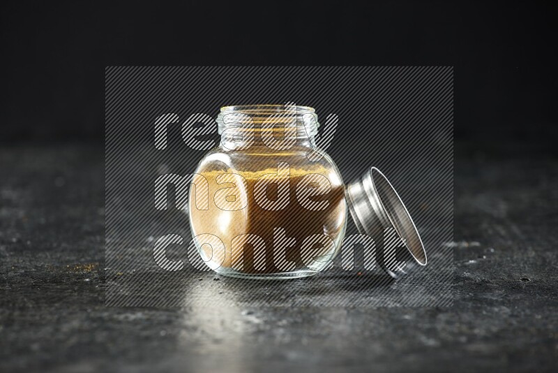 A glass spice jar full of turmeric powder on a textured black flooring