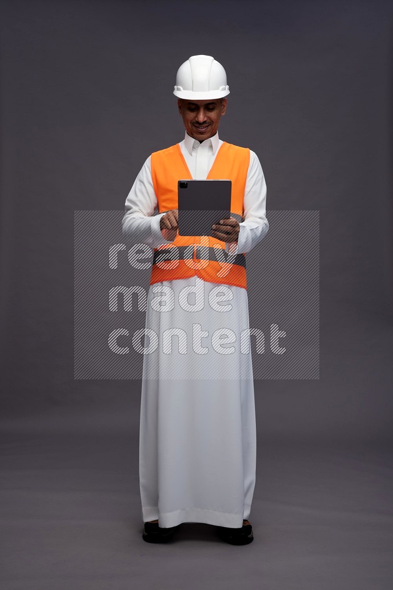 Saudi man wearing thob with engineer vest standing working on tablet on gray background