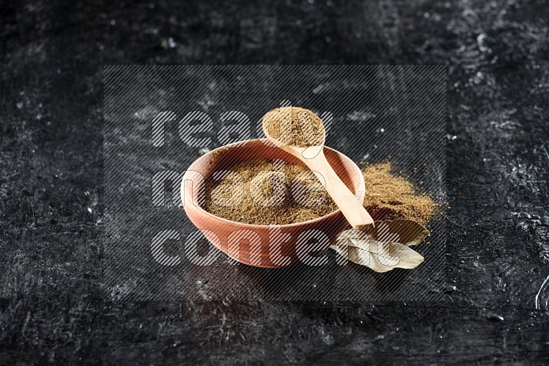 A wooden bowl and spoon full of cumin powder on a textured black flooring