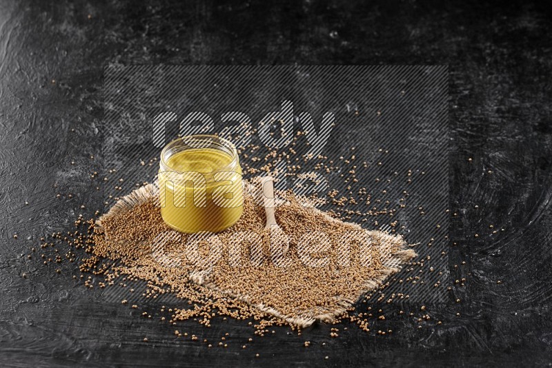 A glass jar full of mustard paste set on a burlap piece with a wooden spoon full of mustard seeds on a textured black flooring