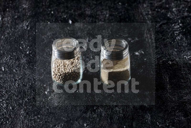 2 Herbal glass jars full of white pepper beads and powder on textured black flooring