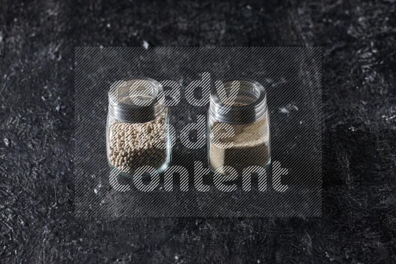 2 Herbal glass jars full of white pepper beads and powder on textured black flooring