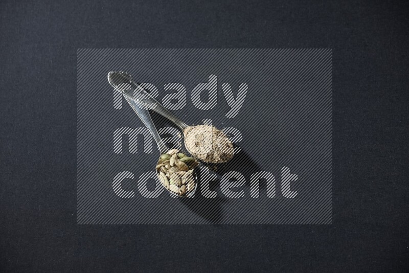 2 Metal spoons full of cardamom powder and cardamom seeds on black flooring