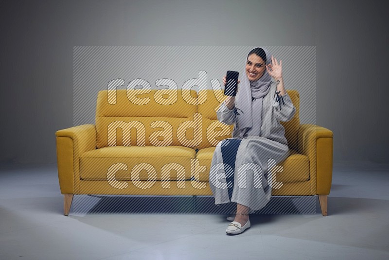 A Saudi woman wearing a light gray Abaya and head scarf setting on a yellow sofa and holding her phone while showing it's screen eye level on a grey background