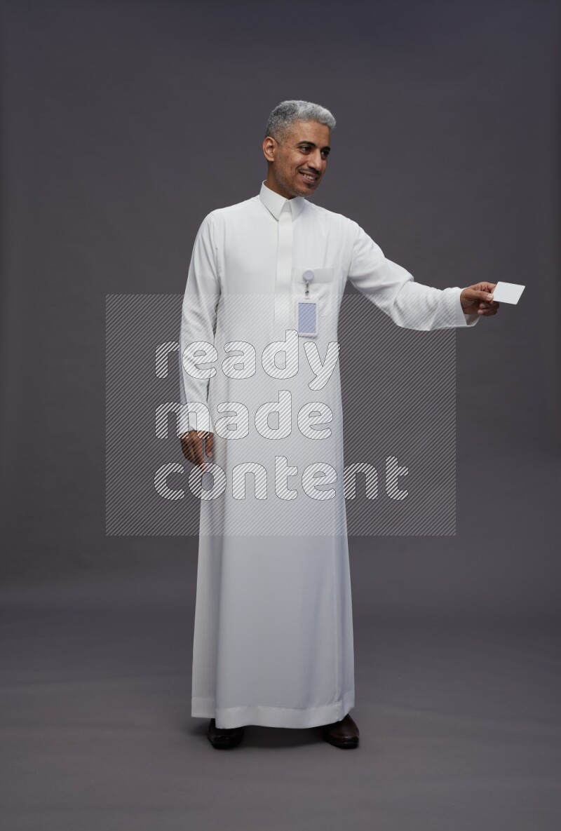 Saudi man wearing thob with pocket employee badge standing holding ATM card on gray background