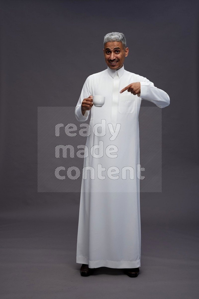 Saudi man wearing thob standing holding mug on gray background