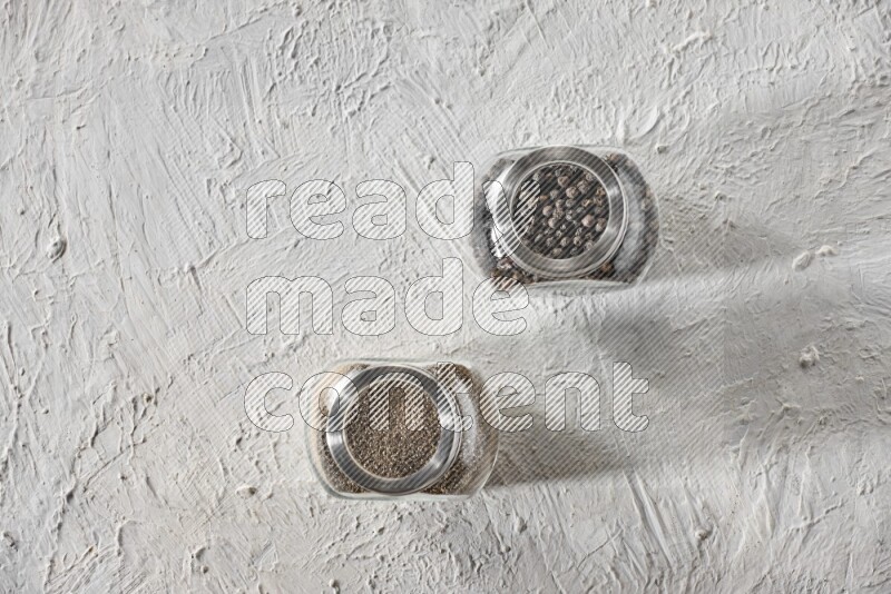 2 glass spice jars full of black pepper and black pepper powder on textured white flooring
